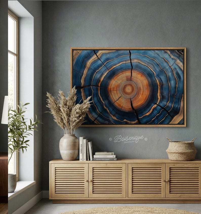 May include: A framed artwork featuring a cross-section of a tree trunk, showcasing concentric rings in shades of blue, orange, and brown. The artwork is displayed above a wooden cabinet with a vase of dried plants, books, and a woven basket.