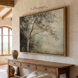 Op de afbeelding: Een groot, ingelijst canvas kunstwerk met een boom in gedempte kleuren groen, bruin en blauw. Het hangt boven een lange, houten console met drie lades. De kamer heeft een rustieke, natuurlijke uitstraling.
