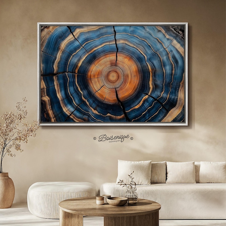 May include: A framed artwork featuring a cross-section of a tree trunk, showcasing concentric rings in shades of blue, orange, and brown. The artwork is mounted in a silver frame and hangs on a neutral-colored wall. The artwork is a decorative piece.
