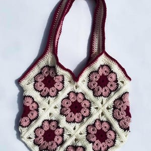 May include: A handmade crochet tote bag featuring a floral pattern. The bag is cream-colored with burgundy and light pink flower motifs. The handles and trim are burgundy. The bag is designed with a square shape and a scalloped top edge.