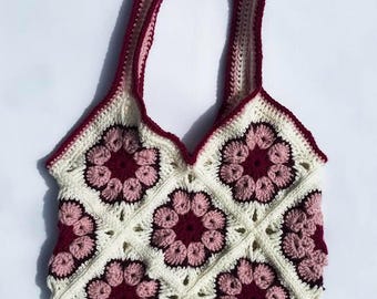 Puede incluir: Bolso tote de crochet hecho a mano con un diseño floral. El bolso es de color crema con motivos florales burdeos y rosa claro. Las asas y el ribete son burdeos. El bolso tiene forma cuadrada y un borde superior festoneado.