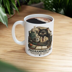 Könnte beinhalten: Ein weißer Keramikbecher mit einer Grafik einer schwarzen Katze mit Brille und Krawatte, die vor einem Computer sitzt und eine Kaffeetasse hält. Der Text lautet "PROGRAMMER CAT DEBUGGED BY COFFEE" mit einem Pfotenabdruck. Gefüllt mit schwarzem Kaffee.