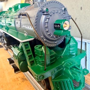 Puede incluir: Un detallado modelo de locomotora en tonos verdes y grises, con el número 515 en la parte delantera. El modelo se encuentra sobre una vía plateada, mostrando su intrincado diseño y estética clásica.