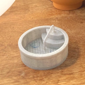 Könnte beinhalten: Ein kleiner, hellgrauer Miniaturbrunnen mit einem mehrstufigen Design steht auf einer Holzoberfläche. Ein terrakottafarbener Topf mit einer grünen Pflanze befindet sich im Hintergrund. Links ist eine große, weiße, runde Lampe.