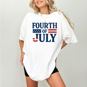 Puede incluir: Camiseta blanca con el texto "FOURTH OF JULY" en azul marino, con un diseño de estrellas y rayas. La camiseta tiene un corte holgado y oversize.