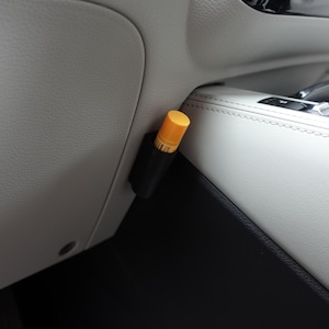 May include: A black and yellow car air freshener is attached to the interior of a vehicle. The air freshener has a yellow top and a black body. The interior of the car is light gray and black.