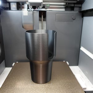 May include: A black, cylindrical cup holder with a handle, freshly printed by a 3D printer. The cup holder is sitting on a textured, tan surface inside the printer. The printer has a dark gray exterior.
