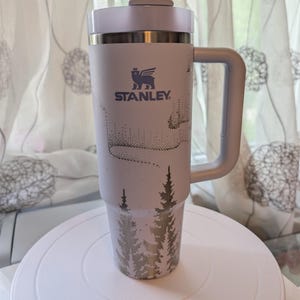 May include: A light lavender Stanley tumbler with a handle, a clear straw, and a lid. The exterior features a dark purple graphic print of a forest of pine trees at the base and a stylized aurora borealis pattern above the Stanley logo and text.