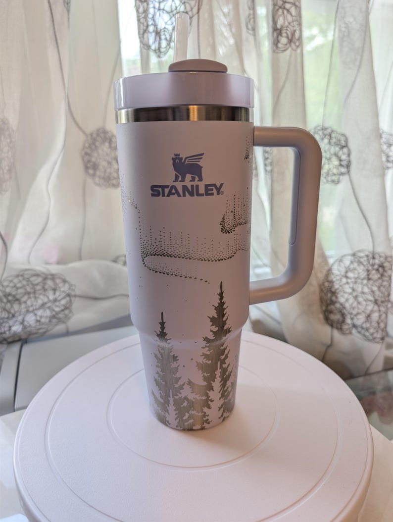 May include: A light lavender Stanley tumbler with a handle, a clear straw, and a lid. The exterior features a dark purple graphic print of a forest of pine trees at the base and a stylized aurora borealis pattern above the Stanley logo and text.