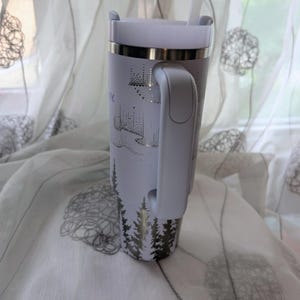 May include: A white insulated tumbler with a handle and a straw, featuring a metallic silver graphic of pine trees at the base and an aurora borealis pattern above. The tumbler has a metallic silver rim and a white lid, standing upright.