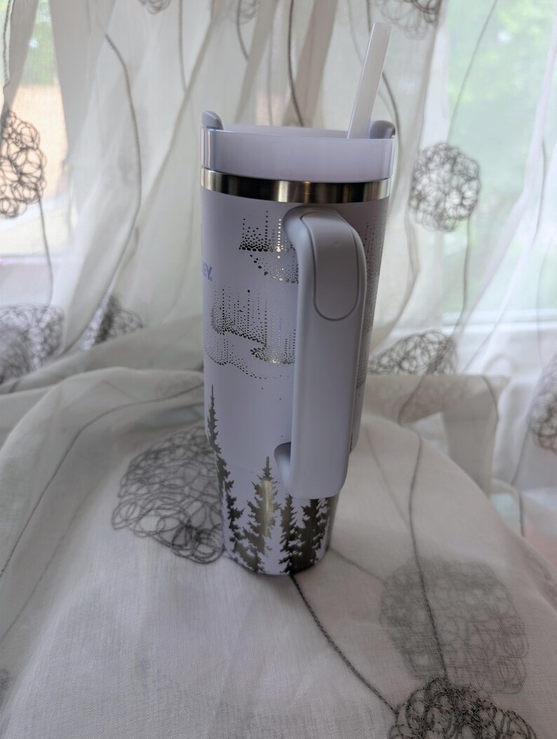 May include: A white insulated tumbler with a handle and a straw, featuring a metallic silver graphic of pine trees at the base and an aurora borealis pattern above. The tumbler has a metallic silver rim and a white lid, standing upright.