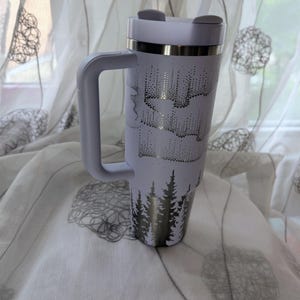 May include: A white insulated tumbler with a handle, a lid, and a straw. The exterior features a dark metallic graphic print of a dense pine forest at the base and an aurora borealis pattern above, wrapping around the cylindrical body.