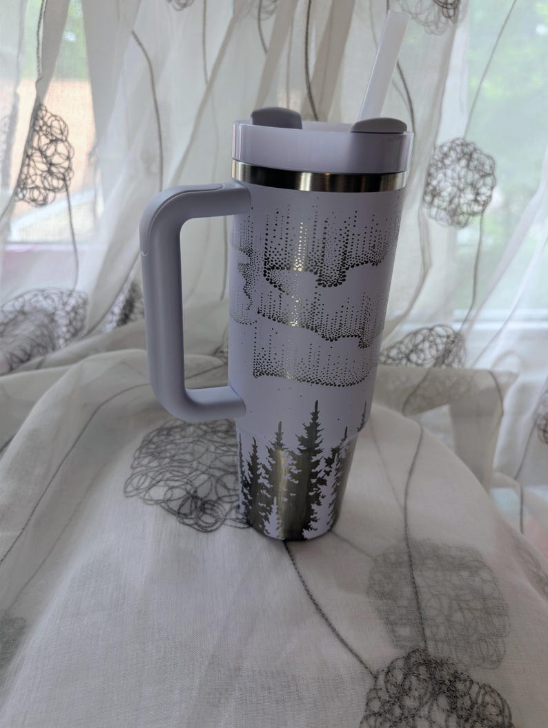 May include: A white insulated tumbler with a handle, a lid, and a straw. The exterior features a dark metallic graphic print of a dense pine forest at the base and an aurora borealis pattern above, wrapping around the cylindrical body.