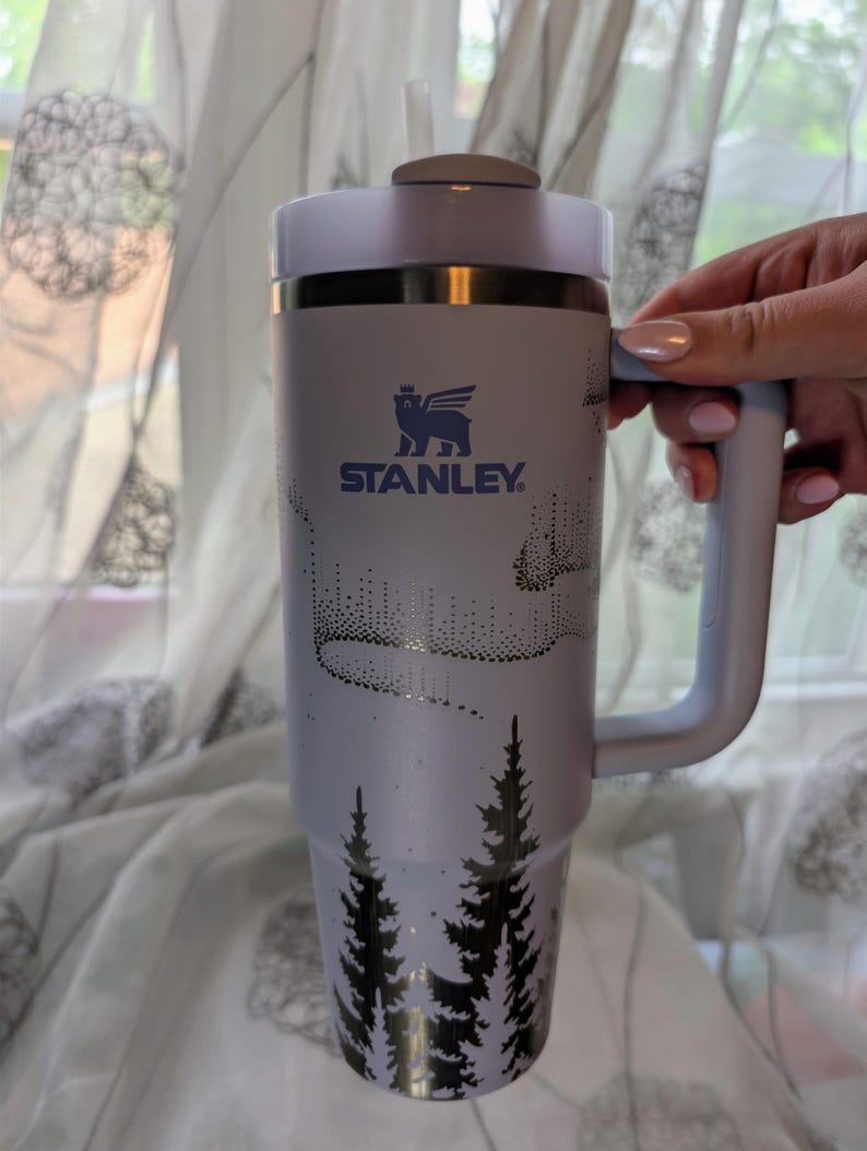 May include: A light purple Stanley tumbler with a handle and a straw, featuring a dark blue logo of a winged bear above the text STANLEY. The cup is decorated with a black graphic print of pine trees at the base and an aurora borealis pattern.