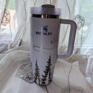 May include: A light purple Stanley tumbler with a handle, a lid, and a straw. The cup features a dark blue Stanley logo and a graphic print of a forest of pine trees at the base with a dotted aurora borealis pattern above the trees on the body.