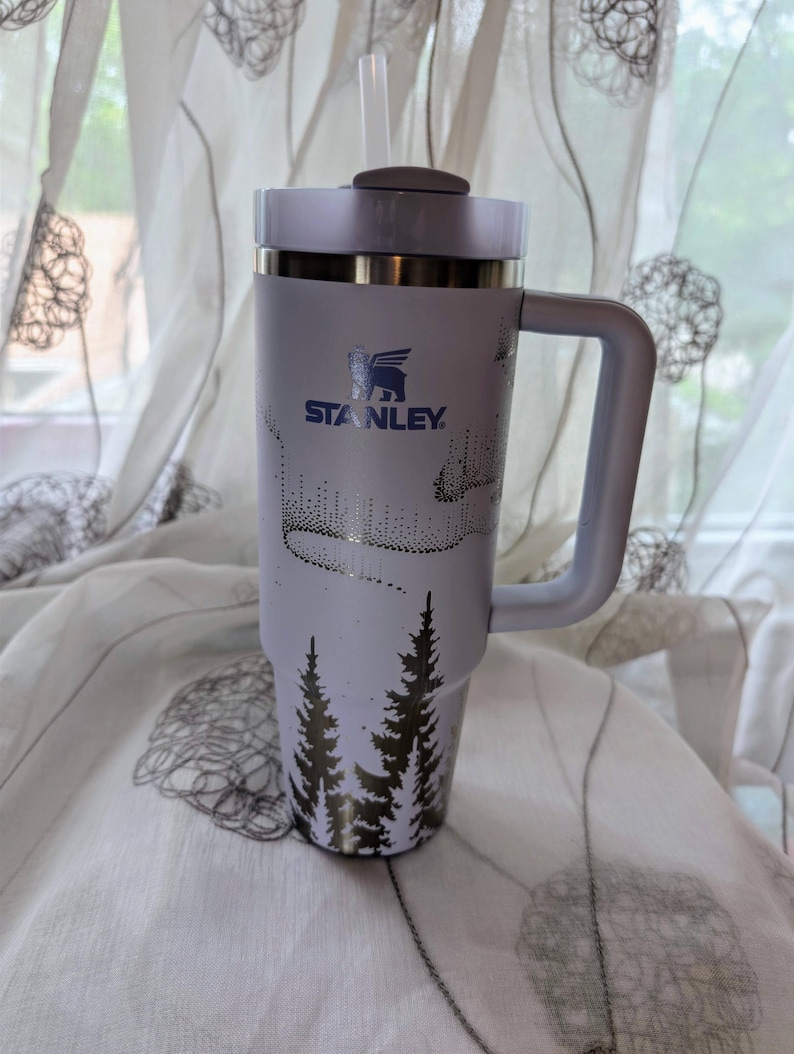 May include: A light purple Stanley tumbler with a handle, a lid, and a straw. The cup features a dark blue Stanley logo and a graphic print of a forest of pine trees at the base with a dotted aurora borealis pattern above the trees on the body.
