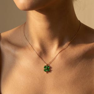 Può includere: Una collana color oro con un ciondolo a forma di quadrifoglio con pietre preziose verdi vibranti. Il quadrifoglio è centrato su una catena delicata, creando un design semplice ma elegante. La collana è indossata intorno al collo.