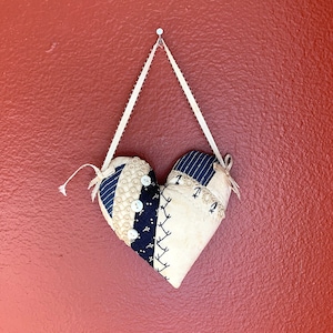 May include: A white and blue patchwork heart-shaped pillow with buttons and stitching details hangs from a white string on a red wall.