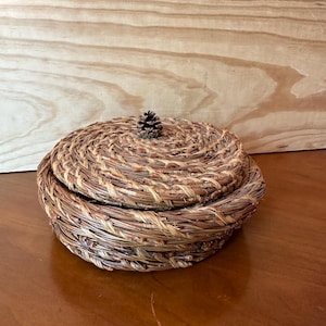 May include: A round woven basket with a lid, made of natural materials like straw or reeds. The lid has a small pine cone on top.