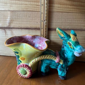 May include: A ceramic donkey-pulled cart planter. The cart is yellow with a pink and red interior, and a red and green wheel. The donkey is teal with yellow and green accents. A ruler is in the background.