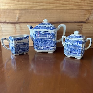 May include: A three-piece blue and white ceramic tea set. The set includes a teapot, a creamer, and a sugar bowl. Each piece features a detailed landscape design. The teapot has a lid and a curved spout. The creamer and sugar bowl have handles.