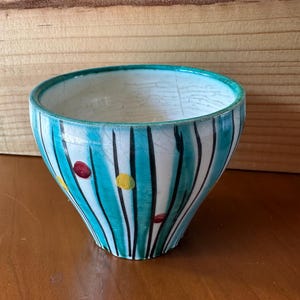 May include: A ceramic bowl with a turquoise rim and vertical stripes in turquoise and black. The bowl has a white background with red and yellow dots. The bowl is on a wooden surface.