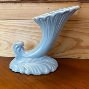 May include: A light blue ceramic vase shaped like a horn with a wavy base.