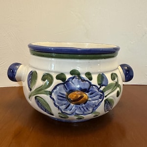 May include: A white ceramic bowl with a blue floral design and two blue handles. The bowl has a blue and green rim and is decorated with blue flowers and green leaves. The bowl is sitting on a brown surface.