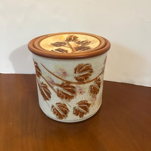 May include: A ceramic jar with a lid, featuring a leaf design. The jar is cylindrical with a cream-colored base and brown leaf patterns. The lid is circular with a brown rim and a central leaf motif. The jar is approximately 6 inches tall.