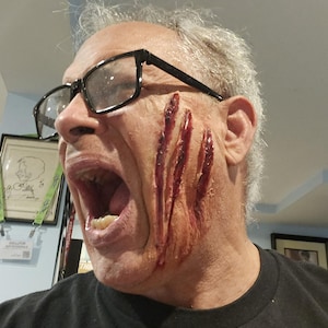 Pode incluir: Uma pessoa com lacerações profundas e sangrentas de aparência realista na bochecha e na mandíbula. A pessoa está usando óculos com armação preta e uma camisa preta, com a boca aberta. A imagem sugere um tema de terror ou efeitos especiais.