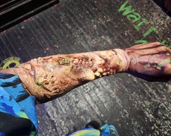 May include: A prosthetic arm with gruesome, realistic details. The skin is a mix of brown, tan, and purple, with exposed flesh and green accents. The arm is positioned against a dark, textured surface, suggesting a horror or special effects theme.