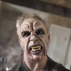 Op de afbeelding: Een close-up van een persoon met zombiemake-up. De huid is bleek met diepe rimpels en aderen. De ogen zijn blauw en de tanden zijn scherp en geel. Bloed druppelt uit de mond. De persoon draagt een zwart shirt.