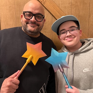 May include: Two people holding star-shaped wands. One wand has a yellow to orange gradient star and a pink handle. The other wand has a blue to pink gradient star and a blue handle. The wands appear to be 3D printed.