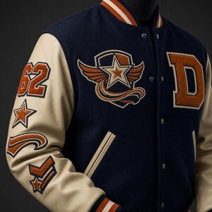 May include: A navy blue varsity jacket with cream-coloured sleeves, featuring orange and white embroidered patches including the number 62, a winged star emblem, and the letter D. The jacket has a snap-button front and striped ribbed collar cuffs.