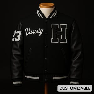 May include: Black varsity jacket with white accents and leather sleeves. Features the number 23 on the left sleeve, the word "Varsity" and a large white "H" on the chest. The jacket has a button-up front and the word "CUSTOMIZABLE" at the bottom.