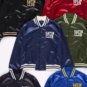 Puede incluir: Chaquetas bomber de satén en negro, rojo, azul marino, verde oliva y azul. Cada chaqueta presenta el texto "SATIN CLUB" en blanco o dorado, con ribetes a rayas en el cuello, puños y dobladillo. Estilo clásico.