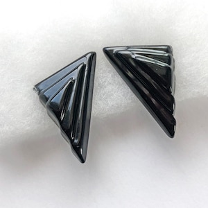 May include: A pair of black earrings with a triangular shape and a glossy, reflective finish. Each earring features a series of three parallel, raised ridges that run diagonally across the surface, creating a stepped, layered appearance.