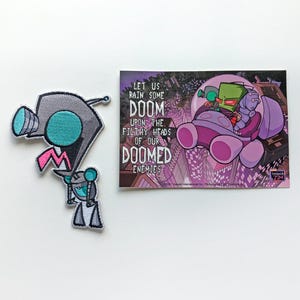 May include: An embroidered patch featuring a gray robot character with a large teal eye and pink mouth, alongside a rectangular sticker depicting a green alien in a purple vehicle with the text: LET US RAIN SOME DOOM UPON THE FILTHY HEADS OF OUR DOOMED ENEMIES.