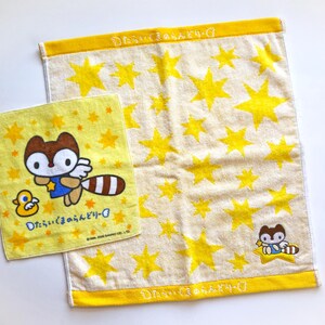 May include: Two yellow and cream-colored towels. One towel features a cartoon raccoon with a duck and Japanese text. The other towel has yellow star patterns and a yellow border with Japanese text. The towels are made of a soft material.