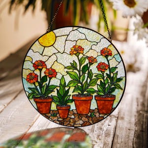 Puede incluir: Vidriera redonda que representa un cielo soleado y flores naranjas en macetas. Las flores están en macetas de terracota y el fondo es un mosaico de colores claros. Una cadena decorativa está adjunta para colgar.
