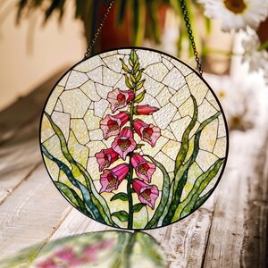 May include: Circular stained glass panel depicting a pink foxglove flower with green leaves, set against a light background. The panel is framed in black and suspended by a chain. The design resembles a mosaic.
