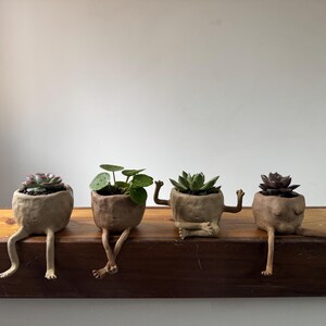以下が含まれることがあります： 人間の足と腕を持つ、手作りの薄茶色の小さな陶器製プランター4つ。それぞれに異なる小さな多肉植物や緑の植物が植えられています。プランターは一列に並べられ、足が縁からぶら下がっている状態です。