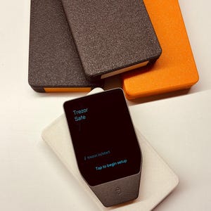 Op de afbeelding: Een verzameling Trezor Safe hardware wallets in verschillende kleuren. De afbeelding toont drie rechthoekige wallets in bruin, donkerbruin en oranje, samen met een apparaat met zwart scherm op een wit kussen. Het scherm toont "Trezor Safe" en "Tap to begin setup."