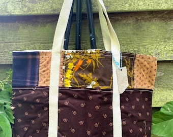 May include: A patchwork tote bag with cream-colored straps. The bag features a variety of fabrics in shades of brown, orange, and blue. The bottom portion of the bag is a dark brown fabric with a floral pattern. The bag is hanging on a black easel.