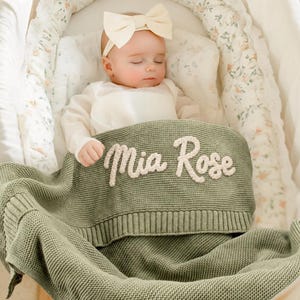 May include: A baby wearing a white long-sleeved shirt and a cream-colored bow headband, lying under a sage green knitted blanket. The blanket features the name Mia Rose embroidered in white, raised, cursive lettering across the top section.
