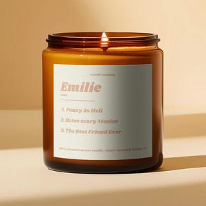 Puede incluir: Una vela de vidrio marrón con una etiqueta blanca que dice "candle mommy Emilie" y enumera tres rasgos de personalidad: "Funny As Hell", "Hates scary Movies" y "The Best Friend Ever". La vela está encendida y la cera se está derritiendo.