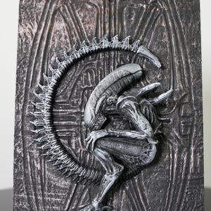 Op de afbeelding: Een gedetailleerd, grijs reliëfsculptuur van een Alien Xenomorph-wezen op een getextureerde, donkergrijze achtergrond. De sculptuur toont het kenmerkende langwerpige hoofd, de ledematen en de staart van het wezen tegen een achtergrond met een getextureerd, industrieel ontwerp.