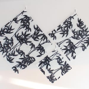May include: Three square fabric cloths of varying sizes, each featuring a white background with a repeating black bamboo leaf pattern. Each cloth includes a small white fabric tag attached to one corner, displaying black text.