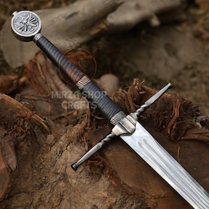 May include: A silver sword with a black leather grip and a silver pommel featuring a winged griffin design. The sword is resting on a wooden surface.