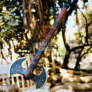 May include: A double-headed axe with a brown leather wrapped handle. The axe head is black and has a hammered finish. The axe is resting on a pile of logs in a wooded area.